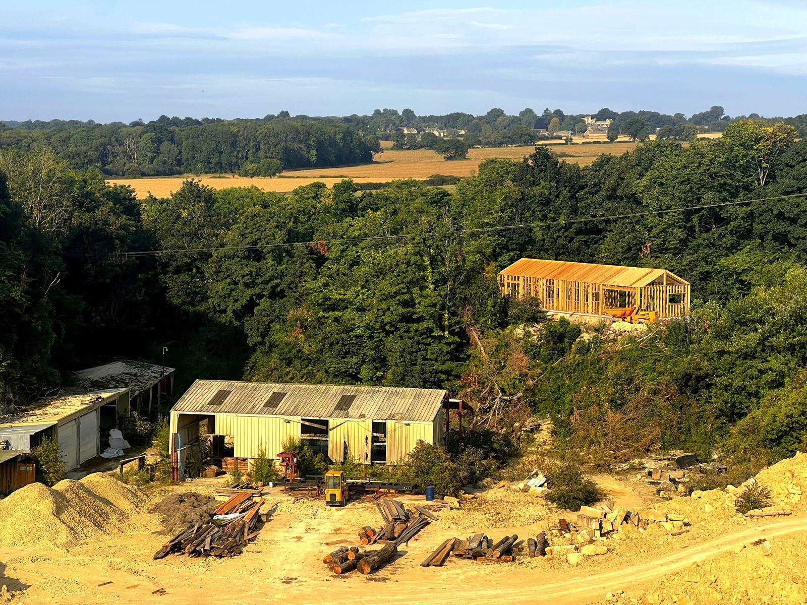 A new building frame rising above a weathered sawmill shed and timber yard in a former limestone quarry, with fields and a village beyond